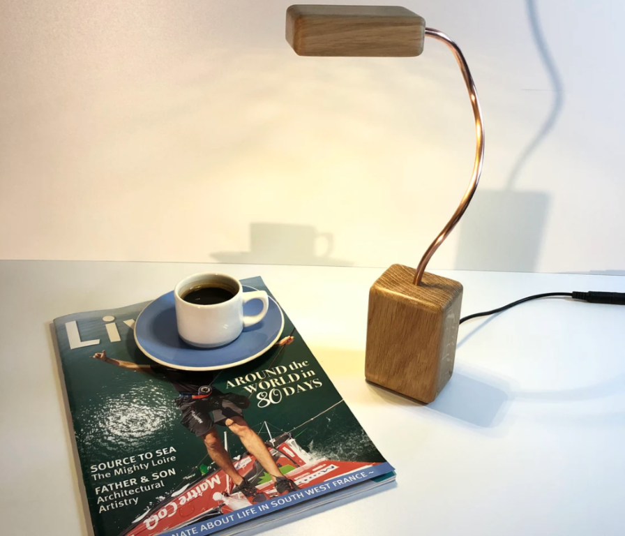 A lamp made from copper and wood is  switched on and lighting up a magazine and white cup filled with coffee. The 3 items are on a white table. Lighting questions